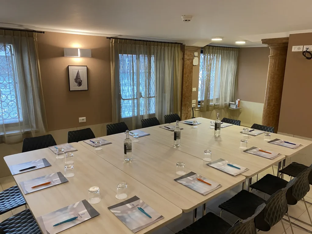 The image shows the hotel meeting room with a large single table in light wood in the center surrounded by black plastic chairs. On the table are folders, pens and bottles of water. From the windows on the left wall you can see the Canal Grande.