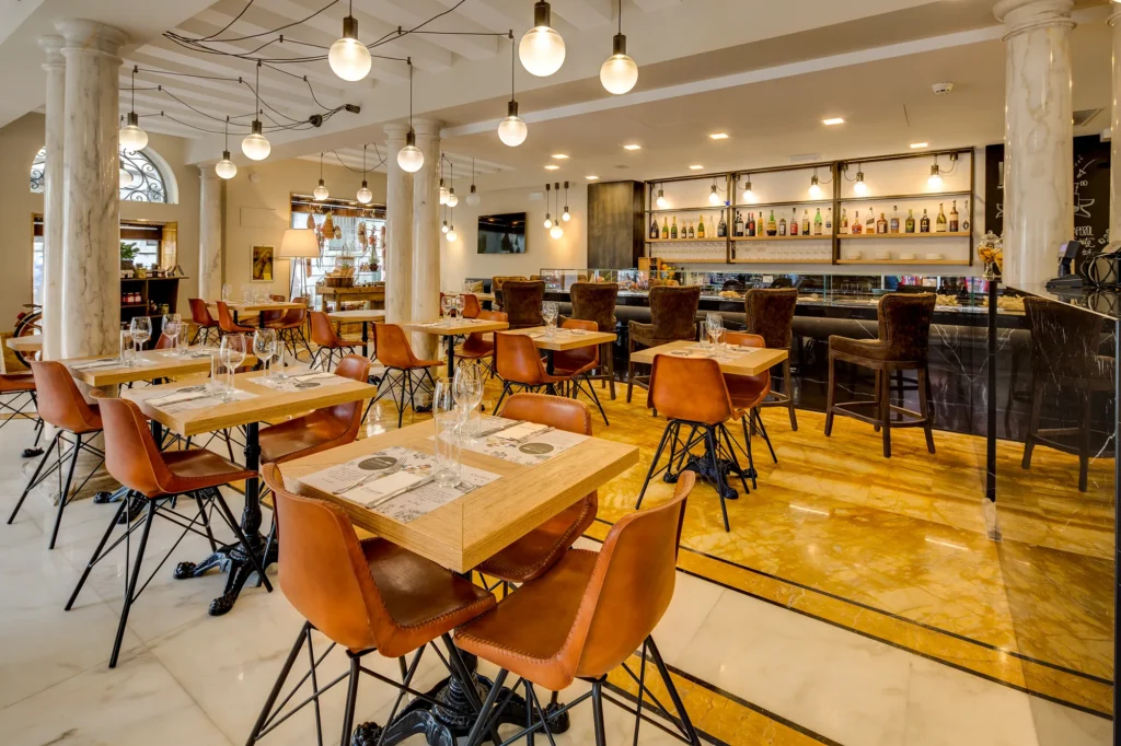 In the picture the Cicchetti Bistrot inside the hotel. The room is occupied by light wood tables with wrought iron legs and orange chairs with black legs. The floor is made of white and yellow marble. On the right is the hotel bar with the counter, velvet stools and shelves of alcohol. The ceiling is made of white beams and round white chandeliers suspended connected to each other by black wires.