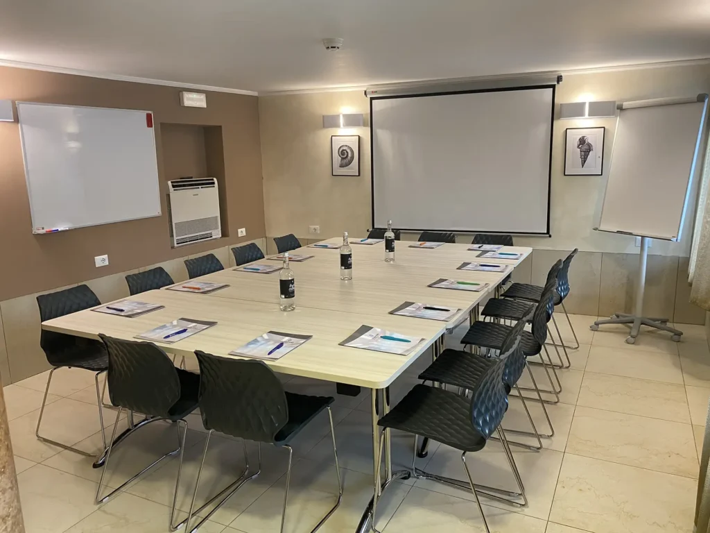 The image shows the hotel meeting room with a large single table in light wood in the center surrounded by black plastic chairs. On the table are folders, pens and bottles of water. On the wall opposite is a projection screen and, next to it, a flip chart.
