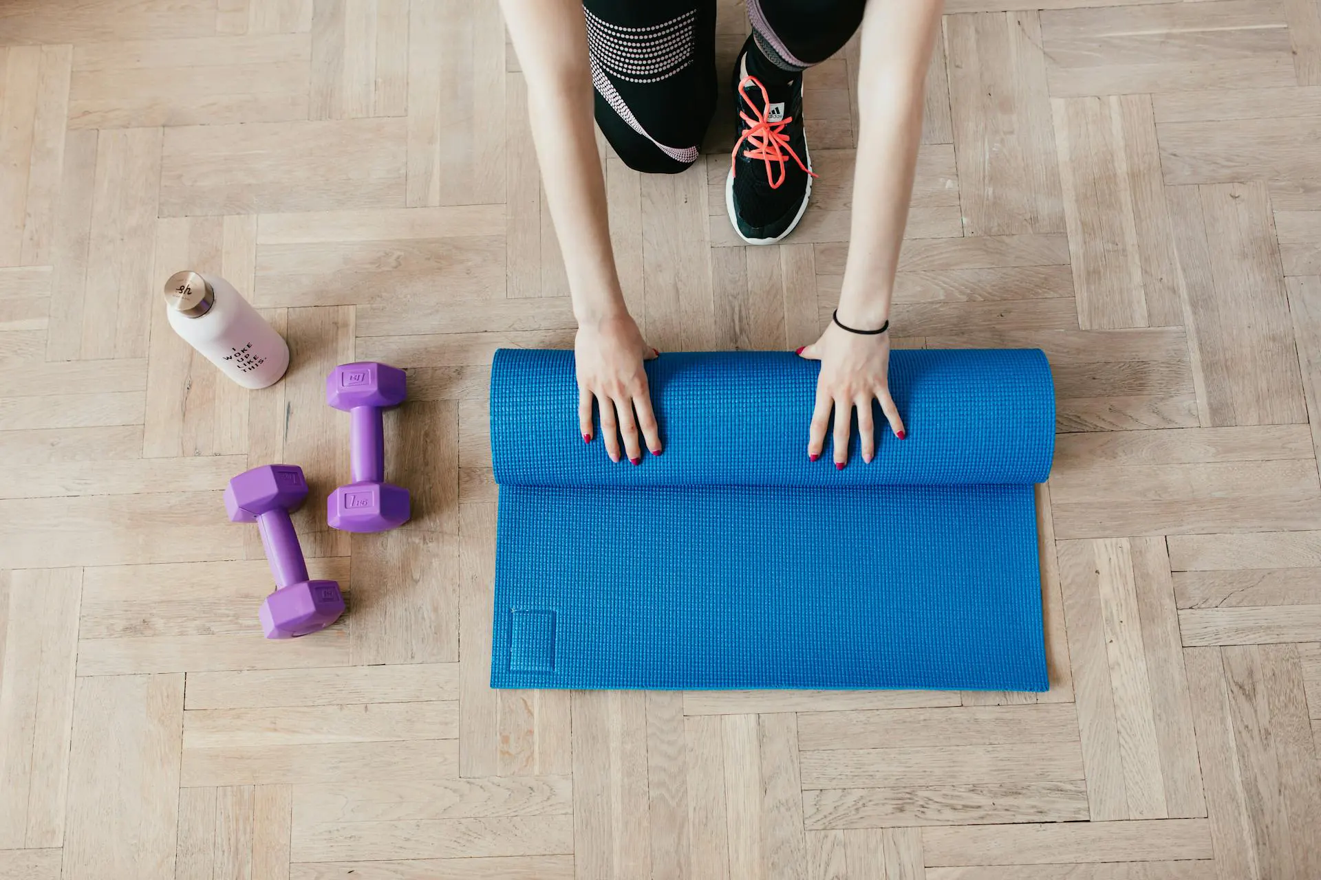 In the picture there is a girl bent over on the ground folding a blue yoga mat. Next to her there are two purple weights and a water bottle